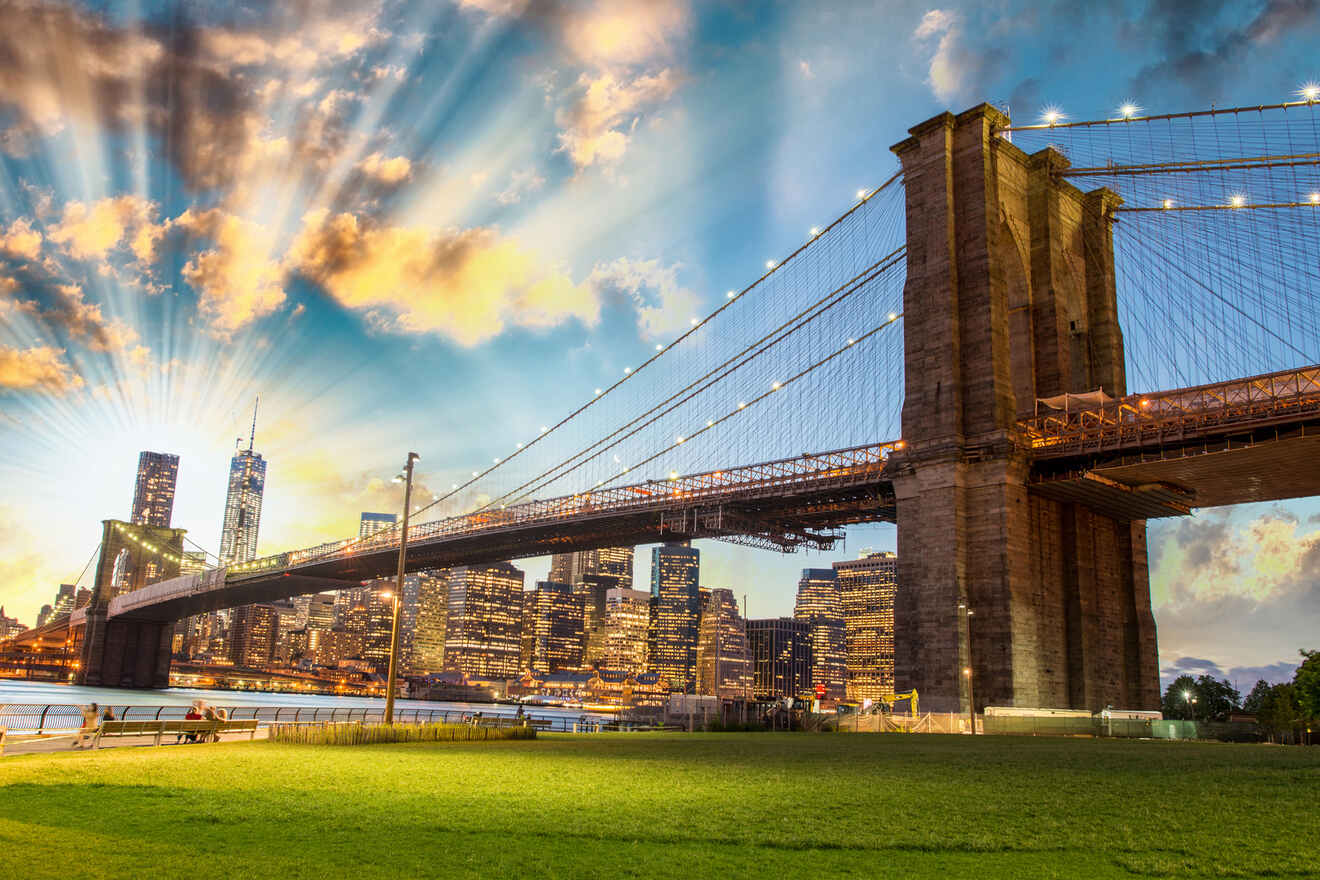 Brooklyn Bridge at sunset with sun rays, a lit cityscape, and a grassy foreground.